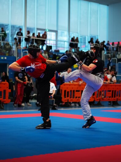 Zwei Kämpfer im Wettkampf, einer in roter und der andere in schwarzer Ausrüstung. Die Atmosphäre ist lebhaft mit Zuschauern im Hintergrund. | © Hütter