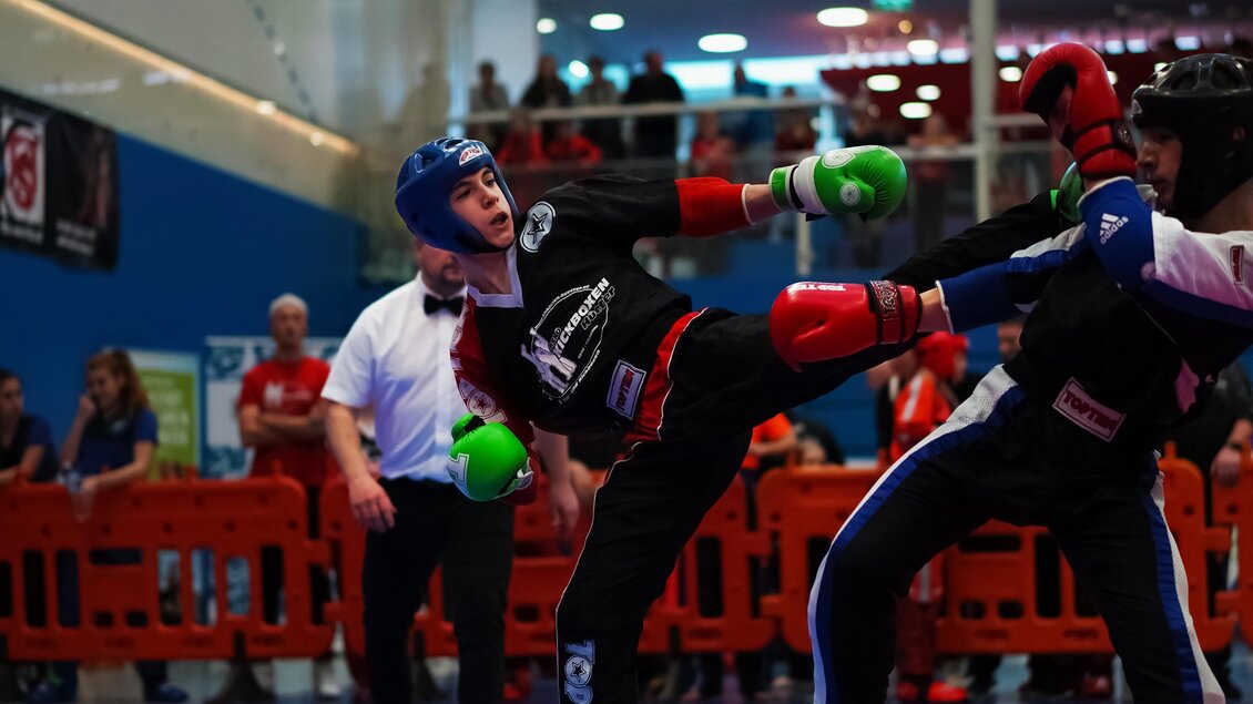 Ein Kampfsportwettbewerb mit Kämpfern in Schutzausrüstung. Einer der Kämpfer führt einen Kick aus, während Zuschauer im Hintergrund zuschauen. | © Hütter