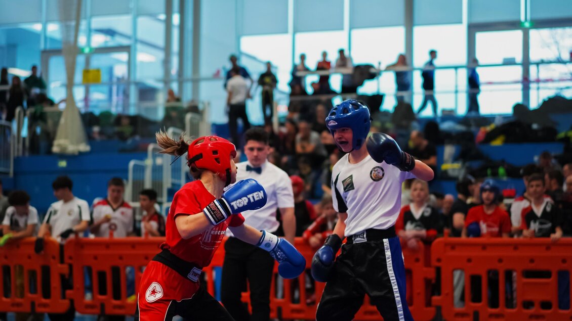 Zwei Kämpfer im Wettkampf, der eine in roter, der andere in blauer Ausrüstung. Im Hintergrund sind Zuschauer und eine Sporthalle sichtbar. | © Hütter