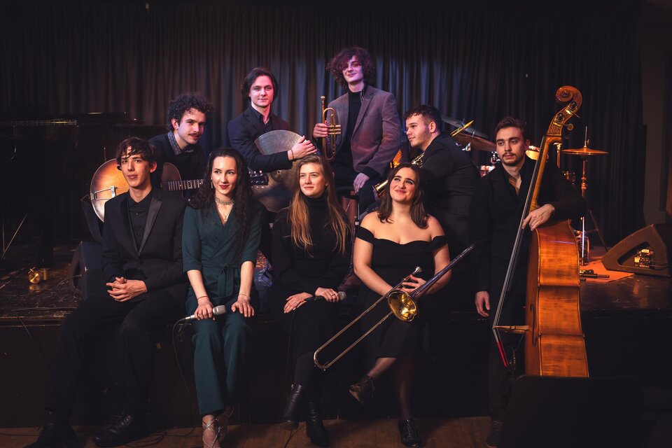 Eine Band aus mehreren Musikern posiert auf einer Bühne. Sie spielen verschiedene Instrumente und tragen schwarze Kleidung. | © KUG Swing Ensemble