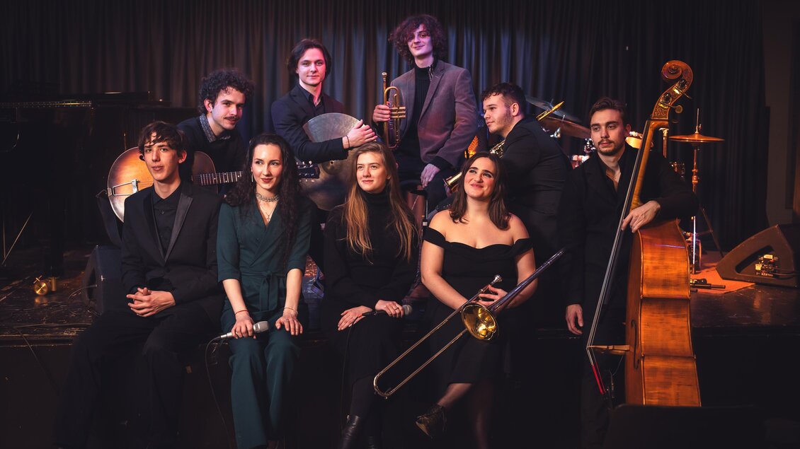 Eine Band aus mehreren Musikern posiert auf einer Bühne. Sie spielen verschiedene Instrumente und tragen schwarze Kleidung. | © KUG Swing Ensemble