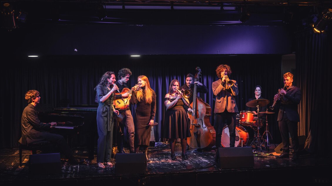 Eine live Musikaufführung mit mehreren Sängern und Musikern auf der Bühne. Im Hintergrund sind Klavier, Kontrabass und Schlagzeug zu sehen. | © KUG Swing Ensemble
