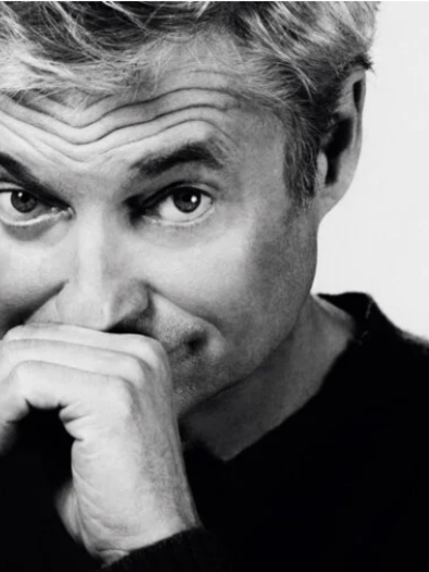 A man smiles mysteriously and holds his hand to his face. In the background, the inscription "DORFER GLEICH" stands out in large, eye-catching yellow. | © Alfred Dorfer