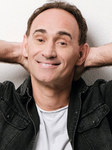 Ein lächelnder Mann entspannt mit verschränkten Armen. Er trägt ein schwarzes Hemd und einen weißen T-Shirt vor einem hellen Hintergrund. | © zur Verfügung gestellt von Plakatoffensive