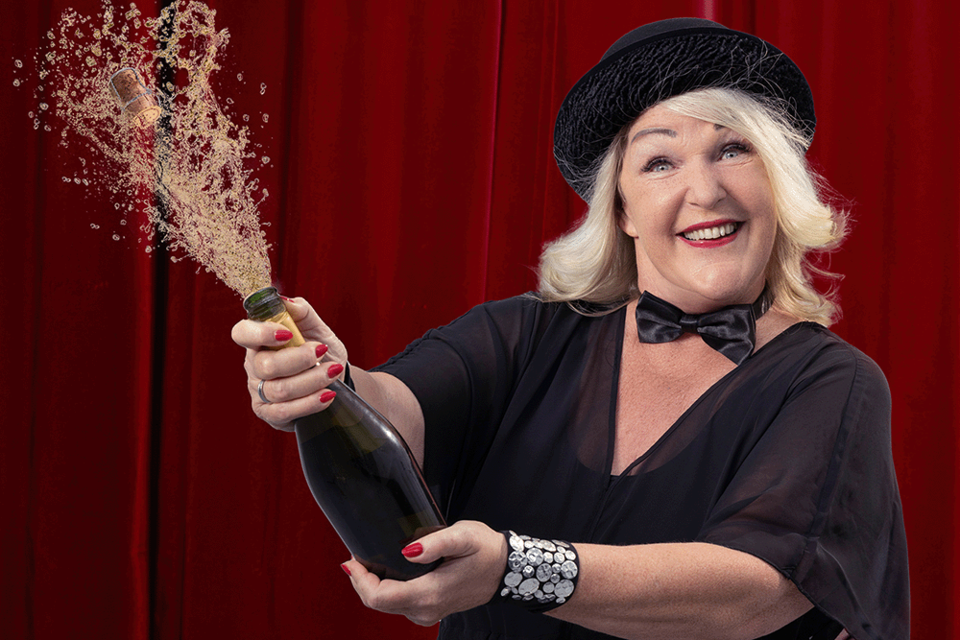 Eine fröhliche Frau mit blonden Haaren sprüht Champagner aus einer Flasche. Sie trägt einen schwarzen Hut und ein elegantes Outfit vor einem roten Hintergrund. | ©  timertl.com