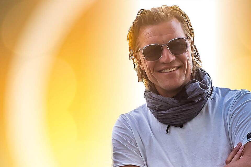 Ein lächelnder Mann mit Sonnenbrille und Schal steht vor einem warmen, goldenen Hintergrund. Er trägt ein weißes T-Shirt und strahlt Freude aus. | © Johann Eder