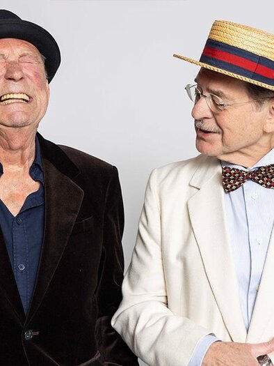 Two older gentlemen smile and look at each other while wearing stylish clothing. One has a black hat and sack suit, the other a white suit with a bow tie and a straw-colored hat. | ©  Xenia Trampusch