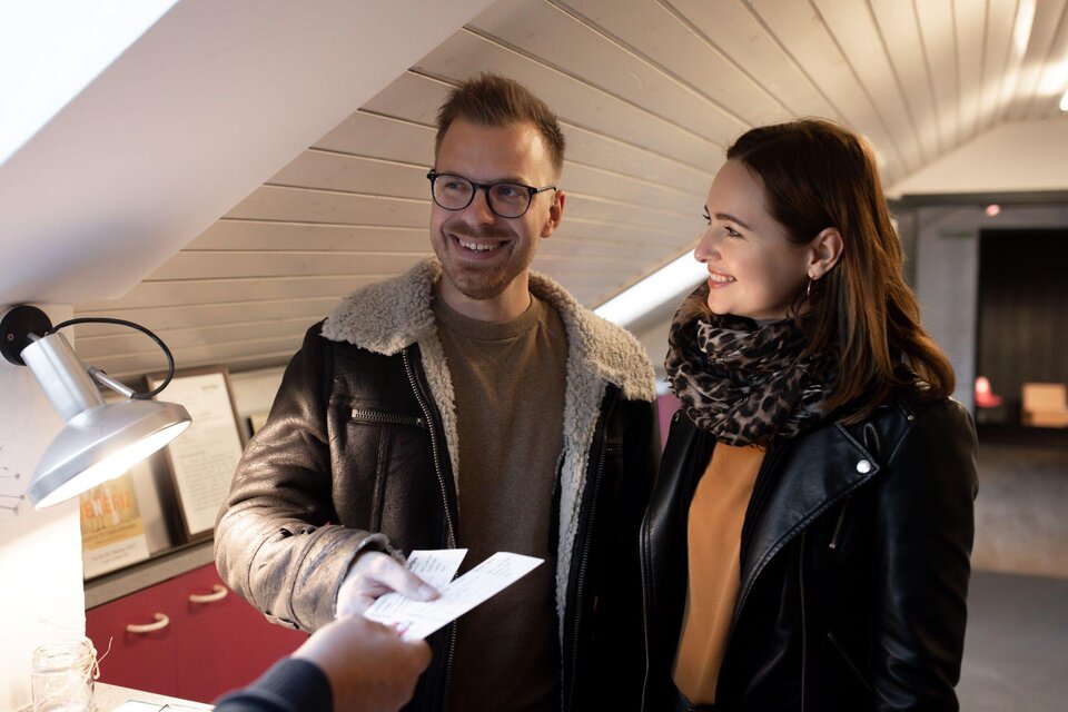 Ein Paar steht in einem Raum und lächelt. Sie halten Papiere in den Händen und scheinen eine Vereinbarung zu treffen. | © TV Südsteiermark - Achromatic Photography