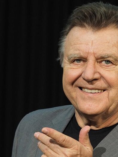 A smiling man in a gray suit who points to himself with one hand. The background is black, providing a clear contrast. | ©  Katrin Werzinger
