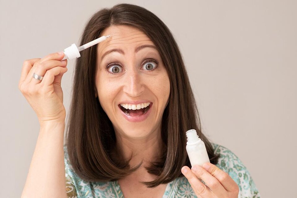 Eine Frau mit langen, braunen Haaren lächelt und hält ein Fläschchen mit einem kosmetischen Produkt in der Hand. Sie zeigt auf ihr Haar, während sie fröhlich in die Kamera blickt. | ©  lynephotography