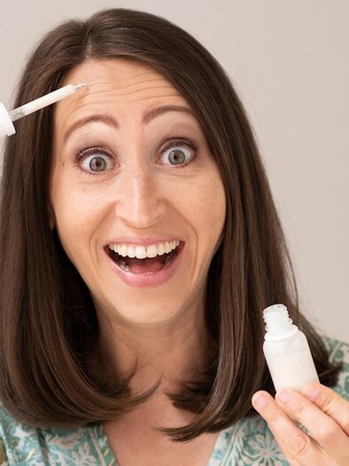 A woman with long, brown hair smiles and holds a small bottle with a cosmetic product in her hand. She points to her hair while happily looking at the camera. | ©  lynephotography