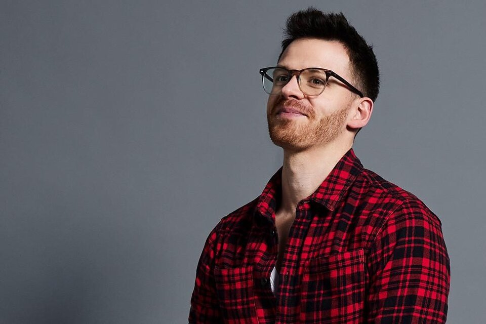 Ein Mann mit Brille und kurzen Haaren trägt ein rot-schwarzes kariertes Hemd. Er schaut lächelnd zur Seite vor einem grauen Hintergrund. | © Andreas Kusy Photography
