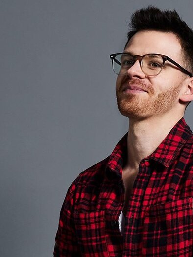 A man with glasses and short hair is wearing a red-and-black checked shirt. He is smiling to the side in front of a gray background. | © Andreas Kusy Photography