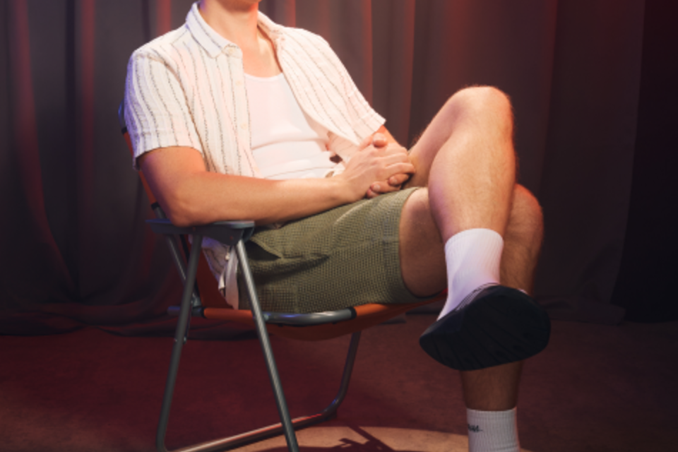 Ein lächelnder Mann sitzt in einem Stuhl und trägt ein offenes Hemd mit einem weißen T-Shirt. Er trägt Socken und Sandalen und hat eine entspannte Haltung. | © KK