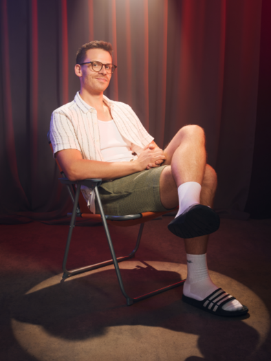 A smiling man is sitting in a chair wearing an open shirt with a white T-shirt. He is wearing socks and sandals and has a relaxed posture. | © KK