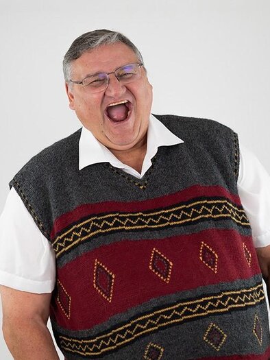 A laughing man is wearing a gray sweater with a colorful pattern. The background is bright and neutral. | © attack