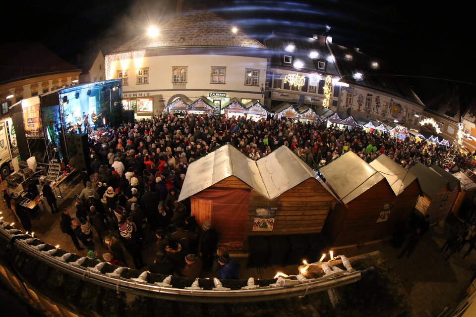 Ein belebter Weihnachtsmarkt bei Nacht mit vielen Menschen und festlicher Beleuchtung. Stände mit traditionellen Holzhütten sind im Vordergrund zu sehen.