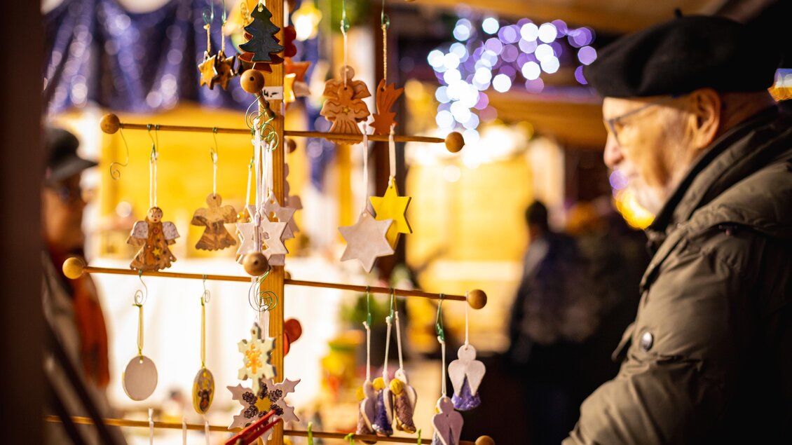 Ein Mann betrachtet dekorative Anhänger auf einem Weihnachtsmarkt. Im Hintergrund leuchten Lichter und Weihnachtsdekoration. | © VANRAY