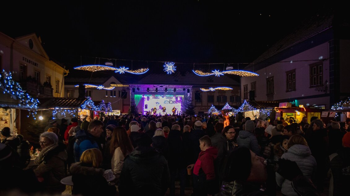 Ein festlicher Platz mit vielen Menschen, beleuchtet durch bunte Lichter. Auf einer Bühne findet eine Veranstaltung statt, umgeben von Ständen und Weihnachtsdekoration.