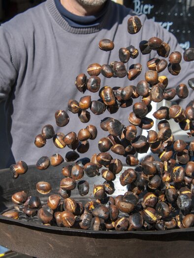 A man is roasting chestnuts in a large pan. The chestnuts fly high and present a vivid picture of the preparation. | © TVB Thermen- & Vulkanland
