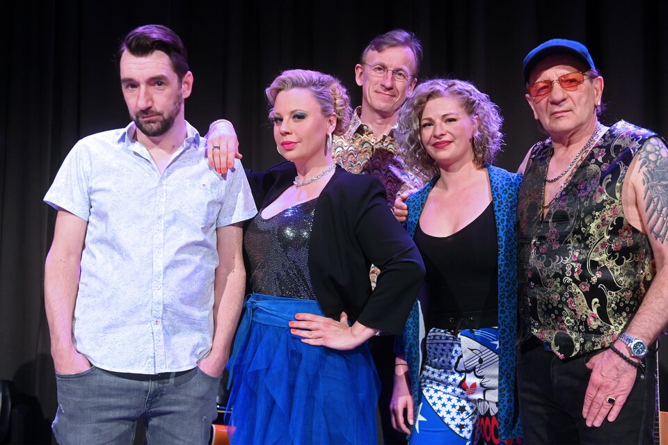 Eine Gruppe von fünf Personen steht lächelnd vor einem dunklen Hintergrund. Jeder hat einen individuellen Kleidungsstil und es scheint eine freundliche Atmosphäre zu herrschen.