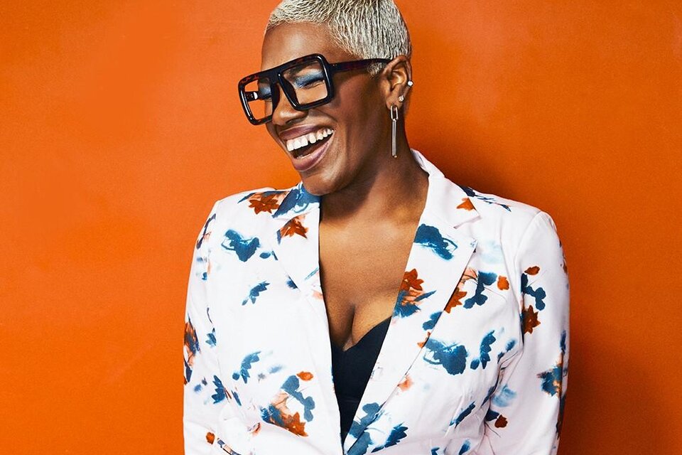 Eine lächelnde Person trägt eine blazer mit buntem Muster und große Brille. Der Hintergrund ist orangefarben. | © ©Peter Lueders