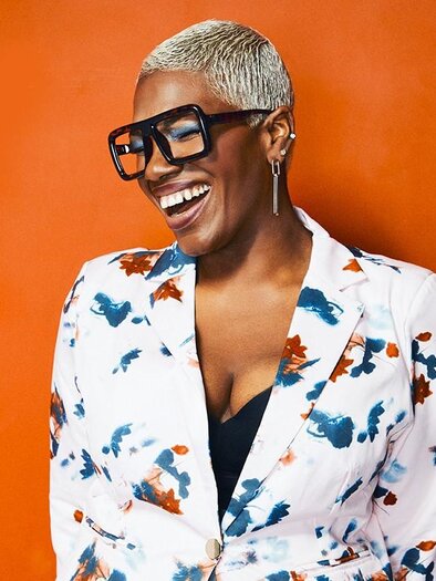 A smiling person is wearing a blazer with a colorful pattern and large glasses. The background is orange. | © ©Peter Lueders
