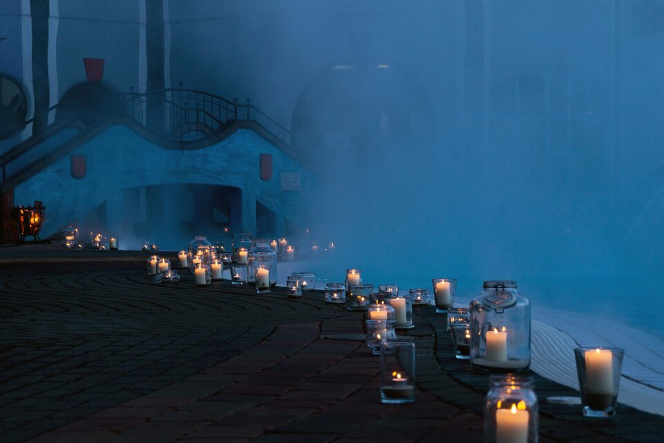 Eine romantische Szenerie mit vielen leuchtenden Kerzen, die einen Weg säumen. Im Hintergrund ist ein Gebäude und leichter Nebel zu sehen. | © Rogner Bad Blumau (C) Hundertwasser Architekturprojekt