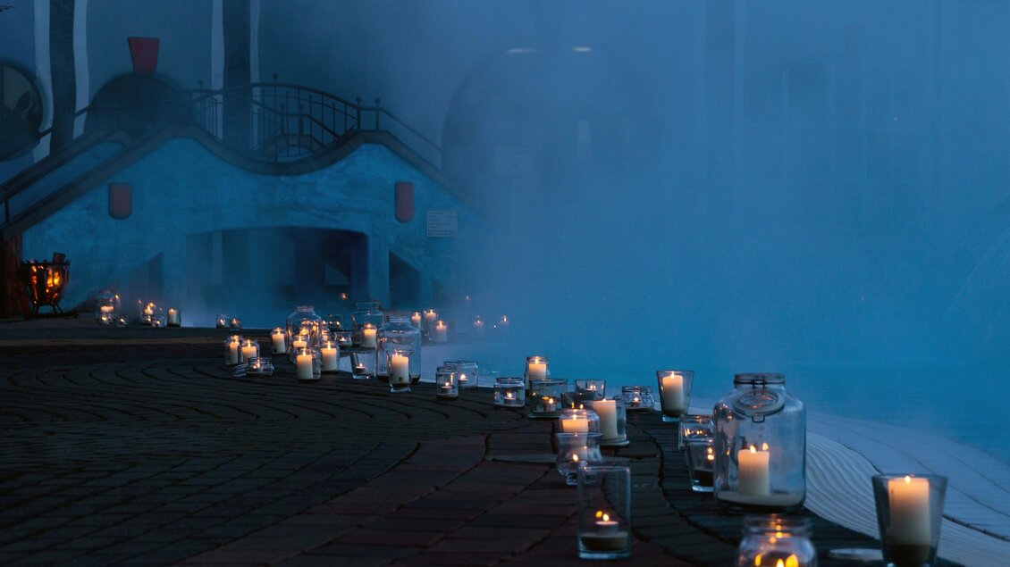Eine romantische Szenerie mit vielen leuchtenden Kerzen, die einen Weg säumen. Im Hintergrund ist ein Gebäude und leichter Nebel zu sehen. | © Rogner Bad Blumau (C) Hundertwasser Architekturprojekt