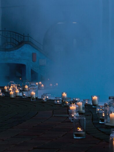 A romantic scene with many glowing candles lining a path. In the background, a building and light mist can be seen. | © Rogner Bad Blumau (C) Hundertwasser Architekturprojekt