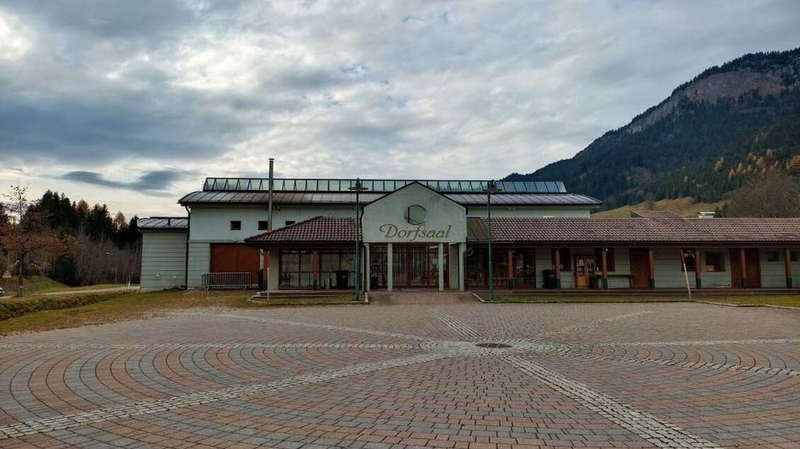 Ein modernes Gebäude mit dem Schriftzug "Dorfsaal" steht vor einer Berglandschaft. Die gepflasterte Fläche vor dem Eingang bildet ein kreisförmiges Muster. | © TVB Ausseerland Salzkammergut_Kolb