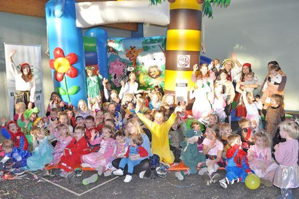 Eine fröhliche Kinderschar in Kostümen versammelt sich um eine Sprunganlage. Die Kinder lächeln und haben Spaß bei der Feier.