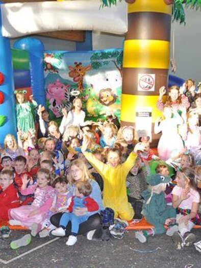 A cheerful group of children in costumes gathers around a jumping facility. The children are smiling and having fun at the party.