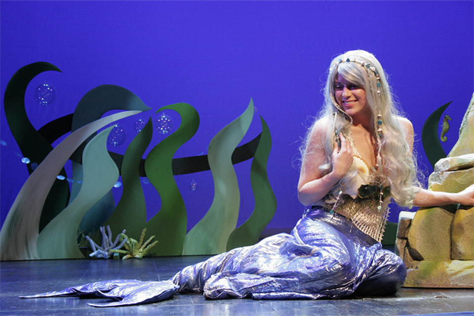 Eine Meerjungfrau sitzt auf der Bühne, umgeben von grünen Pflanzen und Blasen. Sie hat lange, blonde Haare und trägt ein schimmerndes Meerjungfrauenkostüm. | © Theater mit Horizont