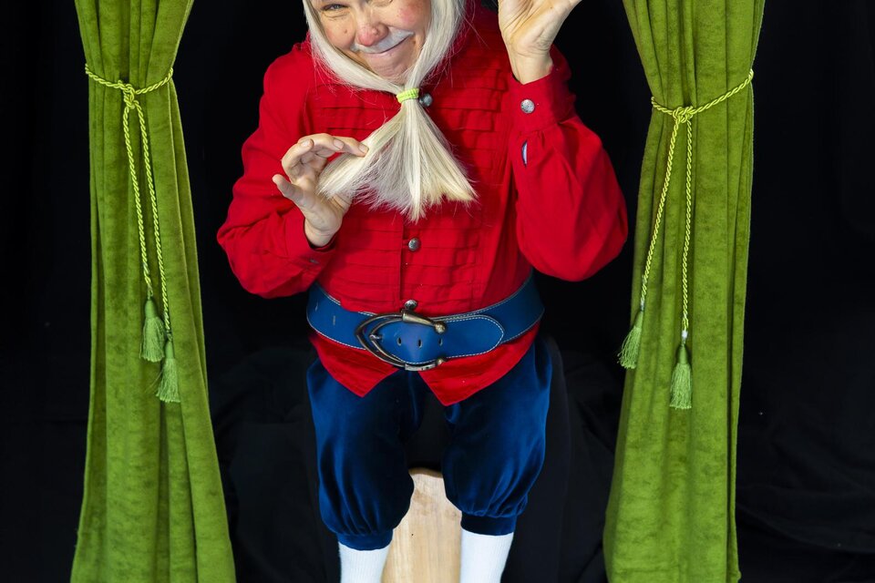 Eine fröhliche Person in einem roten Kostüm mit einer roten Mütze steht auf einem Hocker. Sie posiert unter grünen Vorhängen und hat ein schelmisches Lächeln. | © Dagmar Leis