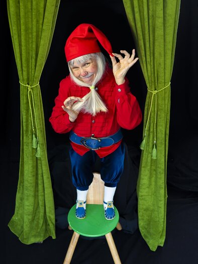 Eine fröhliche Person in einem roten Kostüm mit einer roten Mütze steht auf einem Hocker. Sie posiert unter grünen Vorhängen und hat ein schelmisches Lächeln. | © Dagmar Leis