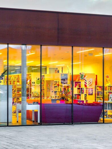 A modern building with large windows and an inviting interior. Shelves with books and cozy seating options are visible. | © Petru Rimovetz
