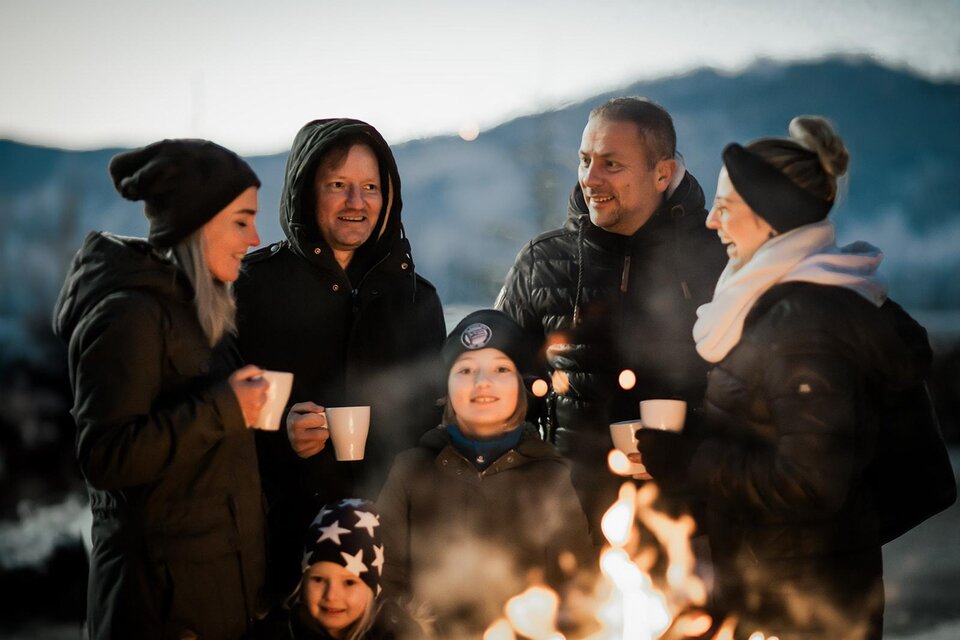 Eine Gruppe von Menschen steht um ein Feuer und genießt warme Getränke. Sie lachen und haben Spaß in einer winterlichen Umgebung. | © TVB Südsteiermark – Achromatic Photography