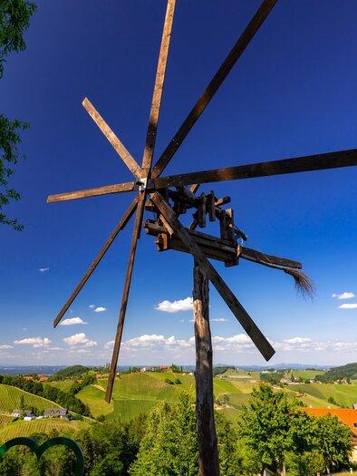 An old windmill stands in a green landscape with gentle hills. The sky is clear and blue, with a few clouds. | © TV Südsteiermark Harry Schiffer