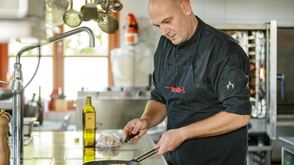 Ein Koch bereitet in einer modernen Küche ein Gericht zu. Er konzentriert sich auf die Pfanne und hat Öl und Zutaten in der Nähe.