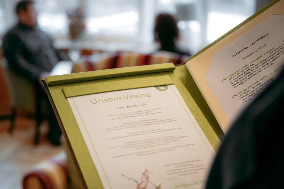 Eine Weinliste in einem Restaurant. Im Hintergrund sind Gäste in einer gemütlichen Umgebung zu sehen. | © Karl Steinegger