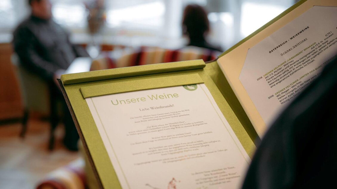 Eine Weinliste in einem Restaurant. Im Hintergrund sind Gäste in einer gemütlichen Umgebung zu sehen. | © Karl Steinegger