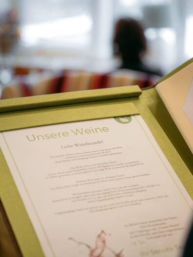 Eine Weinliste in einem Restaurant. Im Hintergrund sind Gäste in einer gemütlichen Umgebung zu sehen. | © Karl Steinegger