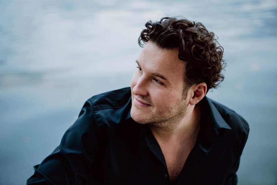 Ein Mann mit lockigem Haar trägt ein schwarzes Hemd und schaut nachdenklich zur Seite. Im Hintergrund spiegelt sich Wasser. | © www.thomas-tatzl.com, Shirley Suarez