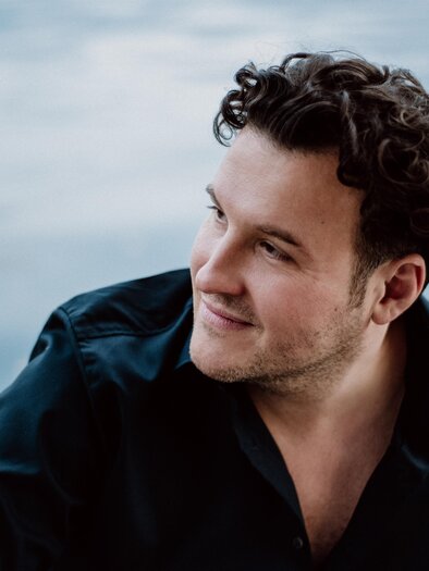 A man with curly hair is wearing a black shirt and is looking thoughtfully to the side. Water is reflected in the background. | © www.thomas-tatzl.com, Shirley Suarez