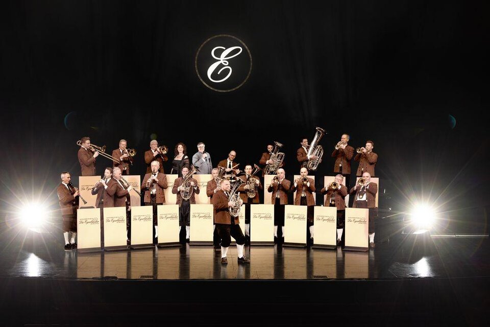 Eine Musikgruppe steht auf einer Bühne und spielt Instrumente. Im Hintergrund leuchtet ein Logo über der Gruppe. | © (c)Paul Gärtne