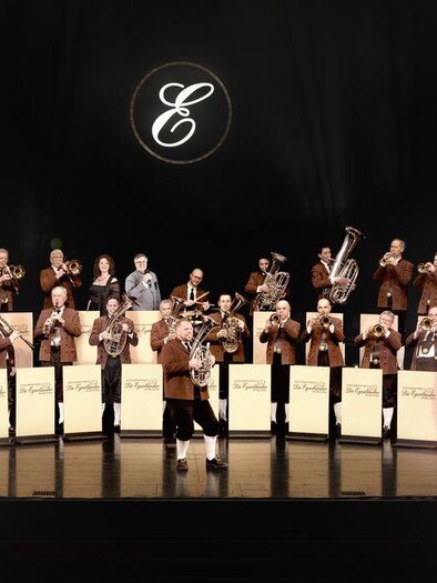 Eine Musikgruppe steht auf einer Bühne und spielt Instrumente. Im Hintergrund leuchtet ein Logo über der Gruppe. | © (c)Paul Gärtne