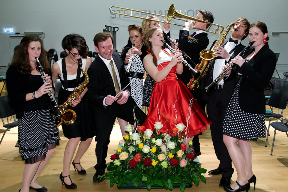 Eine fröhliche Musikgruppe posiert zusammen auf einer Bühne. Die Musiker spielen verschiedene Instrumente und tragen elegante Kleidung, während das zentrale Motiv in einem roten Kleid strahlt. | ©  cp Pictures
