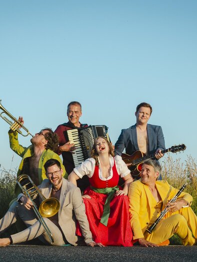 Eine fröhliche Musikgruppe sitzt auf einer Straße, umgeben von hohen Gräsern. Sie spielen verschiedene Instrumente und tragen bunte Kleidung. | © Astrid_Bartl_