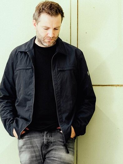 A man in a dark blue jacket is standing relaxed against a light wall. He is looking down and wearing a black t-shirt and jeans. | ©  Philipp Hirtenlehner